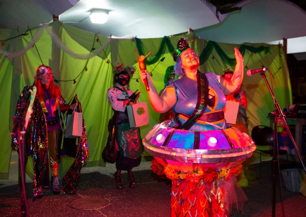 A person in a purple costume with a UFO situated at their hips holds up a slug-shaped trophy with other contestants looking on