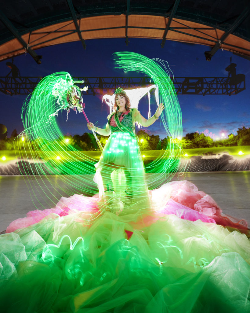 A person in a glowing green dress wields a glowing green staff which has a long-exposure effect giving the impression of it having been swirled in a circle